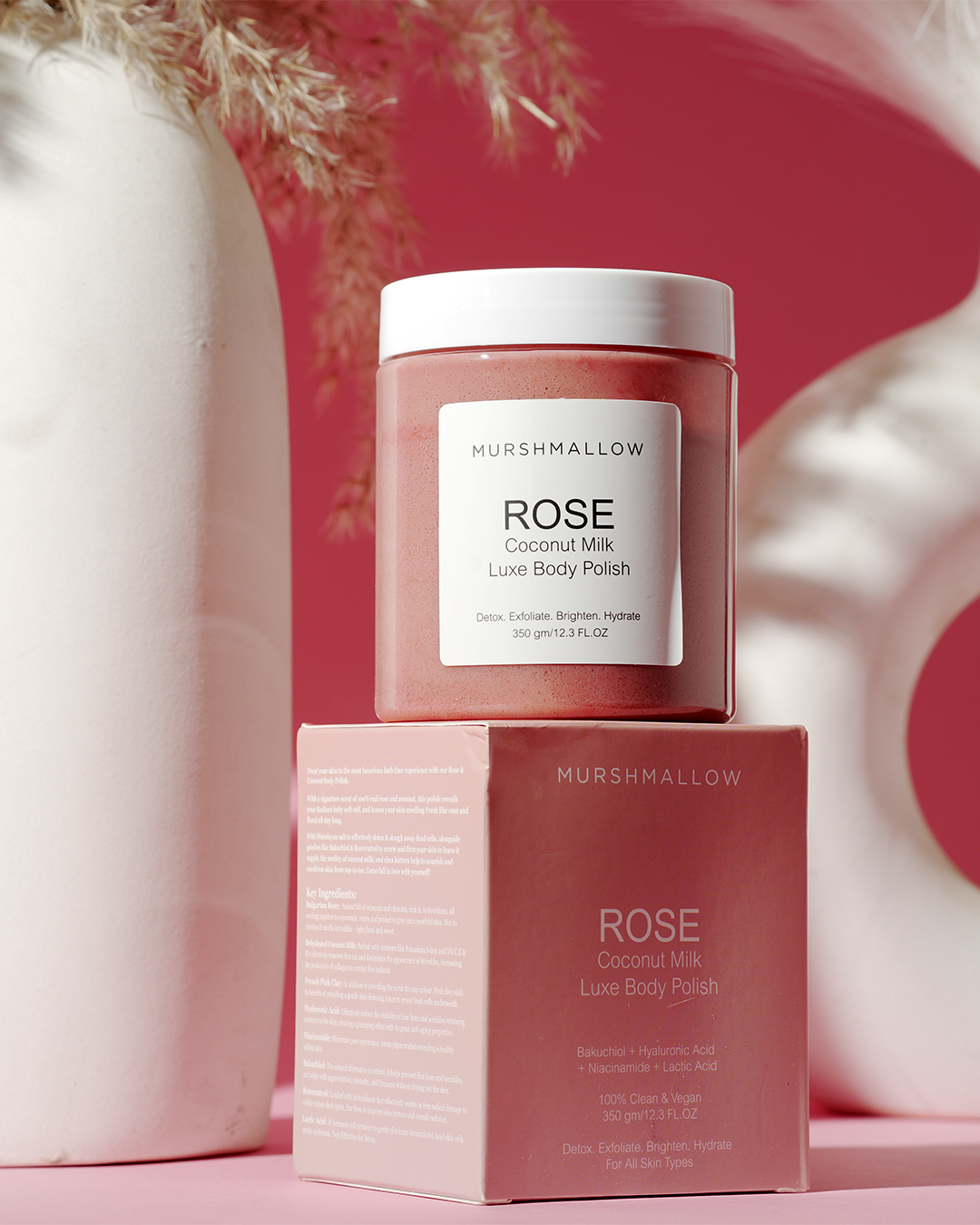 A jar of Murshmallow Rose Coconut Milk Luxe Body Polish stacked on top of its product box, set against a pink background with vases and decorative branches.